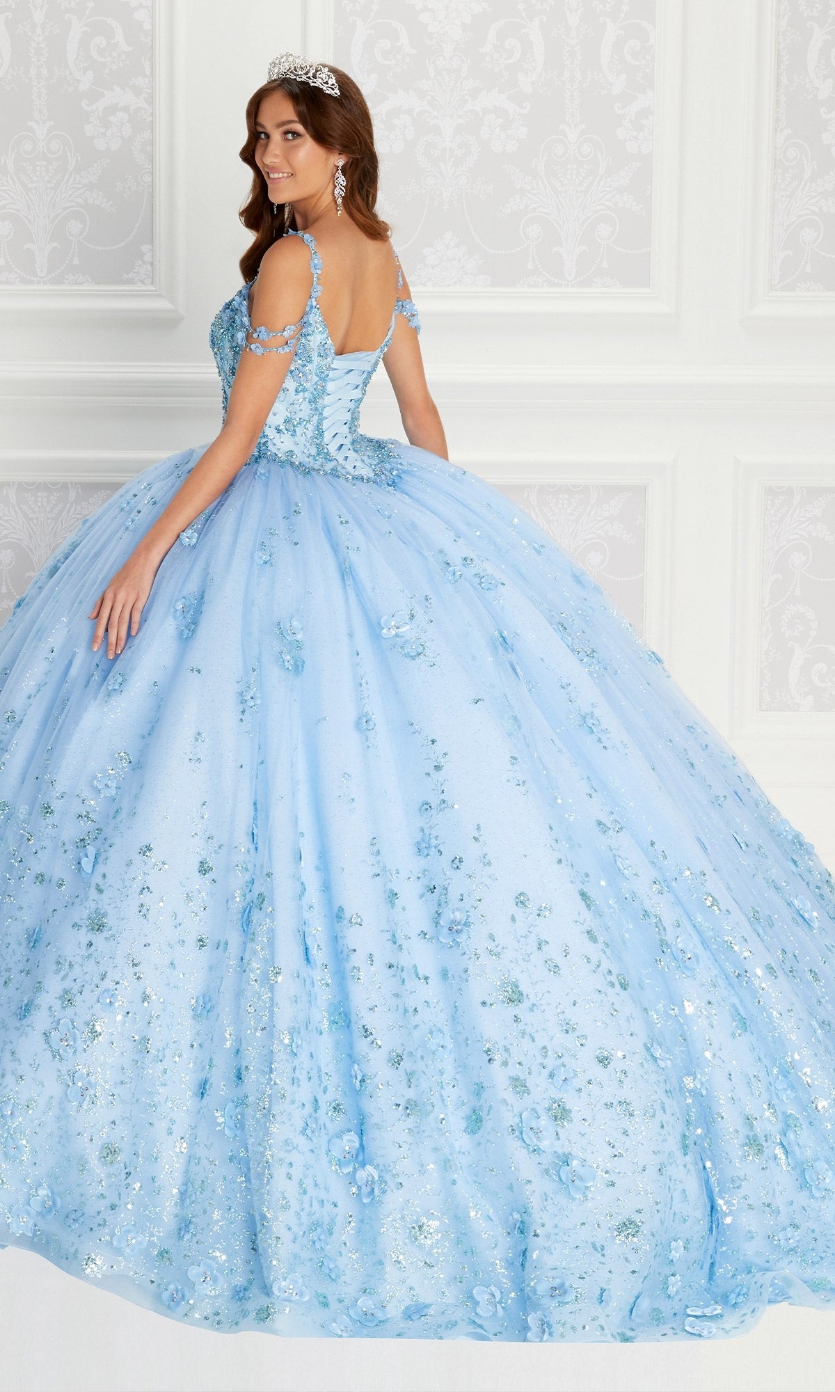 A woman wears the Quinceanera Dress PR22145 By Princesa, a light blue off-the-shoulder gown with floral and rhinestone details, paired with a tiara, as she stands before an ornate white wall.